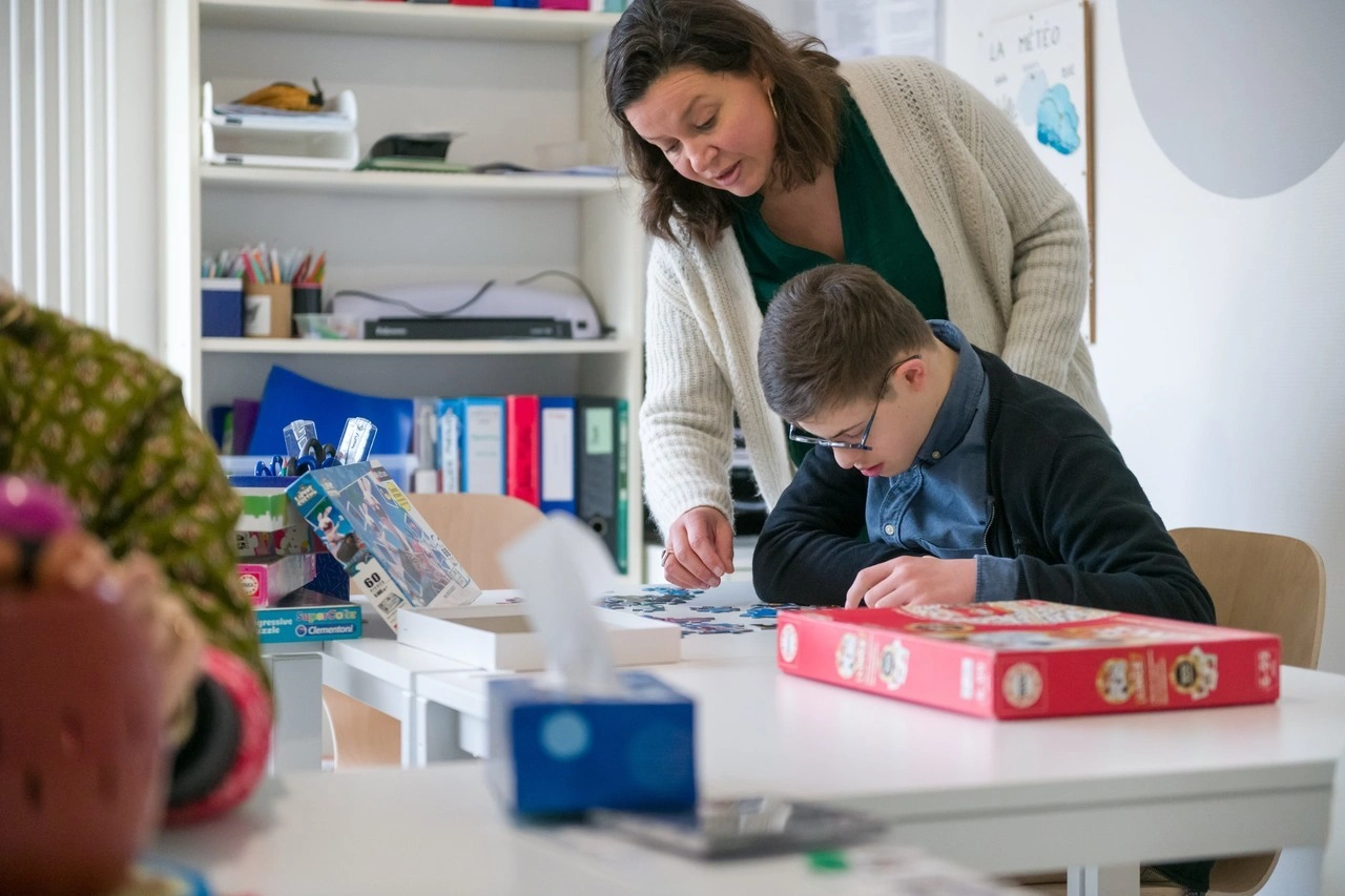 Une femme adulte attentive se penche pour guider un jeune garçon à lunettes alors qu'il se concentre sur l'assemblage d'un puzzle coloré sur une table blanche dans une pièce lumineuse.