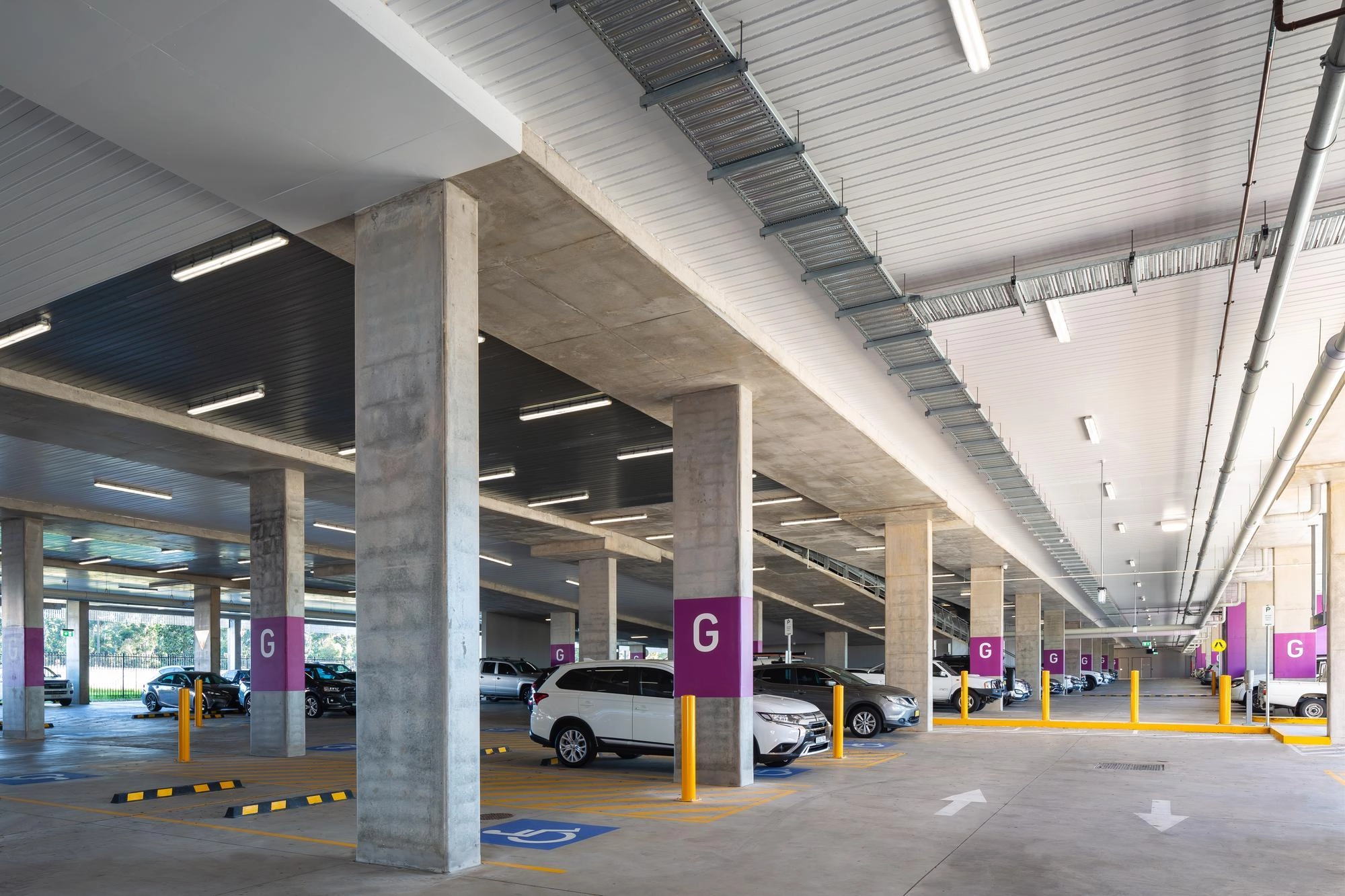 Parking moderne à plusieurs niveaux avec colonnes en béton, panneaux violets 'G' et voitures sous des néons.