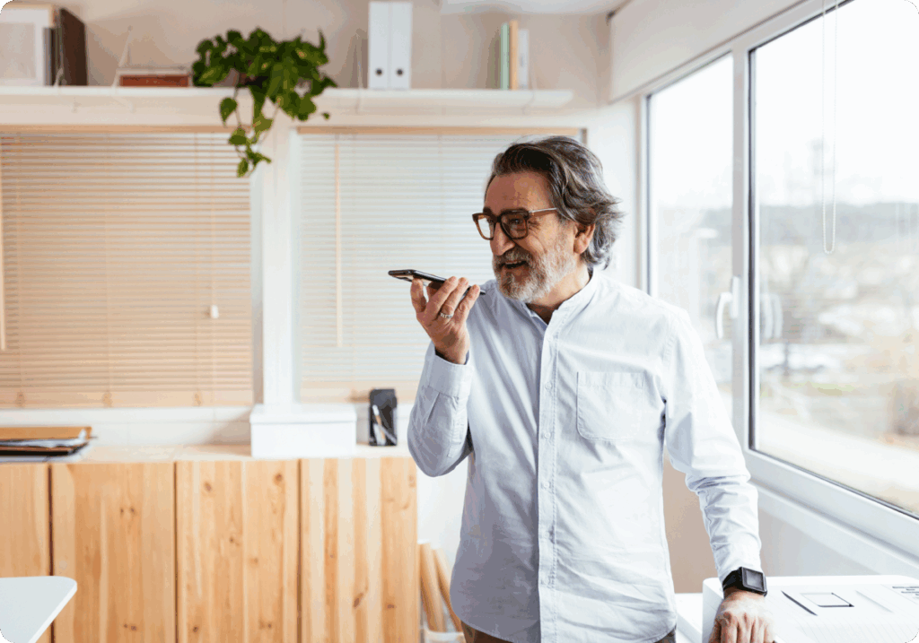 A bearded man wearing glasses holds a smartphone near his mouth in a bright, modern home office, with wooden cabinetry, blinds, shelving and large windows in the background.